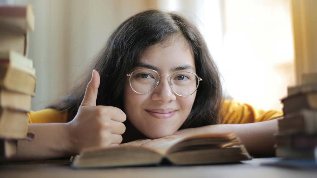 Woman resting her head on a book, smiling and giving a thumbs up