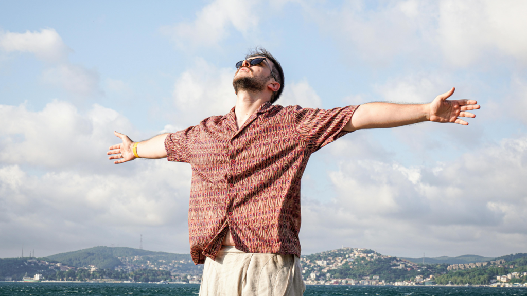 Man standing with open arms beneath a blue sky and next to water