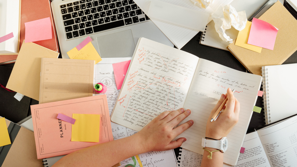 Cluttered desk with laptop, papers and woman writing in a notebook