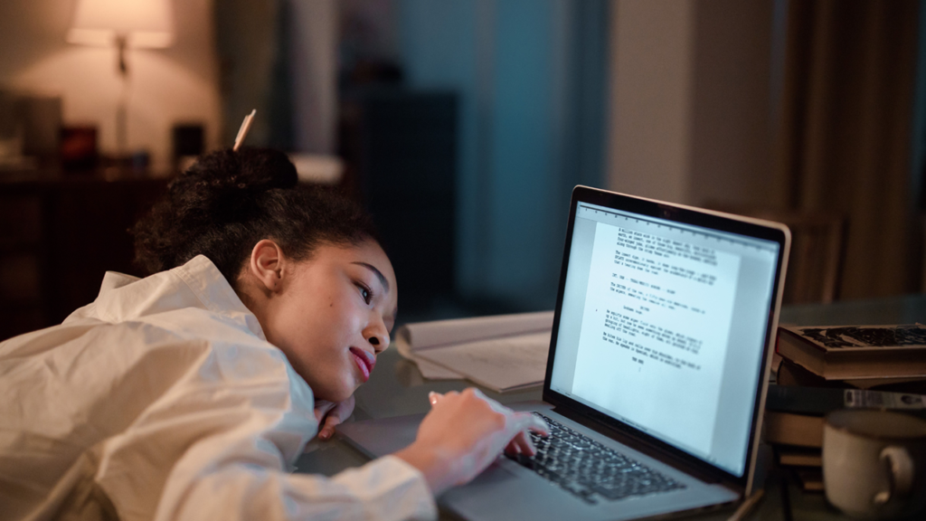 Burned out woman laying on a desk but still trying to type on a laptop